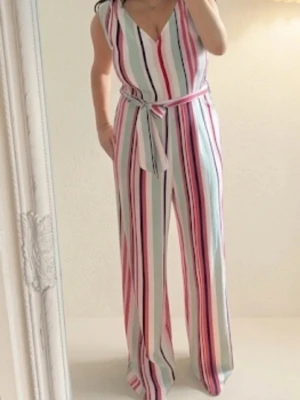 Randig jumpsuit med knytskärp - Snygg jumpsuit med breda ben och vertikala ränder i rosa, vitt, grönt och svart. Djup v-ringning och matchande knytskärp i midjan. Ärmlös modell i ett mjukt, lätt tyg som ger en avslappnad och trendig look.