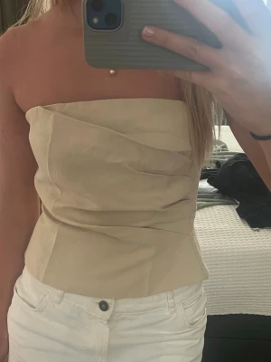 Beige axelbandslös tubtopp - Snygg beige tubtopp med draperad design och figurnära passform. Toppen är axelbandslös och har en stilren look som passar perfekt till både jeans och kjol. Materialet känns mjukt och lätt mot huden.