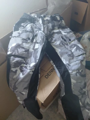 Camo Mc byxor med svarta detaljer - Säljer ett par grå camo Mc byxor med svarta förstärkningar längs benen och stora fickor med lock. Byxorna har bälteshällor och justerbar midja. Perfekta för kalla dagar när du vill hålla stilen och vara bekväm. Sitter skydd i dom för knä rygg 