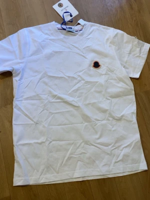 Vit t-shirt från Moncler - Säljer en klassisk vit t-shirt från Moncler med rund hals och diskret logotyp på bröstet. T-shirten har korta ärmar och är tillverkad i mjuk bomull. Perfekt för en clean och stilren look.