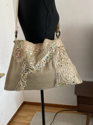 Beige axelväska med spets och paljetter - Unik beige axelväska i tyg med bred axelrem dekorerad med spets. Väskan har detaljer i virkad spets och färgglada paljetter på framsidan, vilket ger en bohemisk och kreativ vibe. Perfekt för dig som gillar vintage och handgjorda detaljer.
