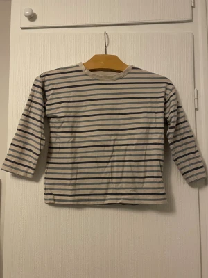 Randig långärmad t-shirt Lindex Kids - Säljer en klassisk långärmad t-shirt från Lindex Kids i storlek 110. Tröjan är vit med tunna blå ränder och har en enkel, rak passform. Tillverkad i mjuk bomull som är skön mot huden. Perfekt för en avslappnad och stilren look.