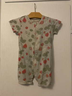 Kortärmad pyjamas med jordgubbstryck Lindex - Supersöt kortärmad pyjamas från Lindex i ljusgrått med glada jordgubbar och gröna blad som mönster. Dragkedja framtill för enkel påklädning. Perfekt för varma dagar. Mjuk och skön bomullskvalitet.
