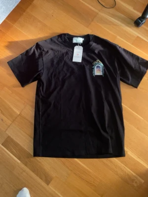Svart t-shirt från Casablanca - Säljer en svart t-shirt från Casablanca med tryck på bröstet och klassisk rund hals. T-shirten är kortärmad och har en stilren look med diskret logga och grafiskt motiv framtill. Perfekt för dig som gillar streetwear och vill ha något trendigt.