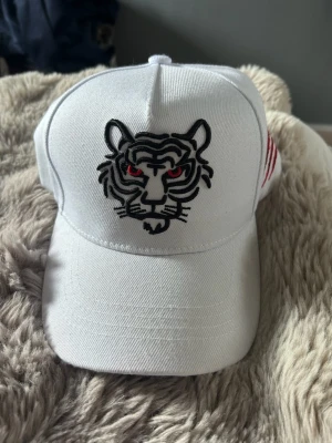 Vit keps med tigerbrodyr - Säljer en vit keps med broderad tiger på framsidan i svart och röda detaljer för ögonen. På sidan finns röda och svarta klösmärken broderade. Kepsen har böjd skärm och är tillverkad i bomullstyg. Perfekt för dig som vill sticka ut med en cool accessoar.