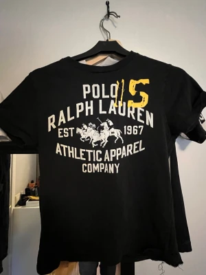 Svart Polo Ralph Lauren t-shirt - Snygg svart t-shirt från Polo Ralph Lauren med stort vitt tryck och gul detalj framtill. Klassisk rund hals och korta ärmar. Tillverkad i mjuk bomull som känns skön mot huden. Perfekt för en sportig och avslappnad look.