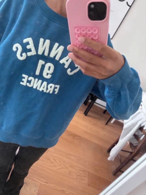 Blå hoodie med Cannes-tryck - Blå hoodie med vit text 'CANNES 91 FRANCE' på bröstet. Oversized passform med ribbade muddar vid ärmslut och nederkant. Mjuk och skön bomullsblandning, perfekt för chill dagar eller när du vill ha en avslappnad look.