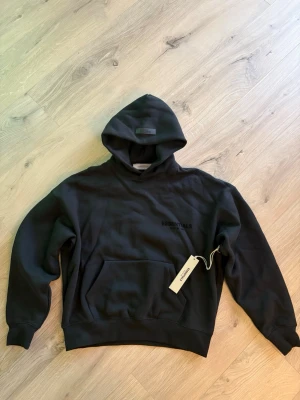 Svart Essentials Fear of God hoodie - Svart hoodie från Essentials Fear of God,köpte fel storlek men kunde inte lämna tillbaka. Diskret logga på bröstet och stor logga på ryggen. Mjuk bomullskänsla och ribbade muddar vid ärmslut och midja. Perfekt för en avslappnad streetwear-look.