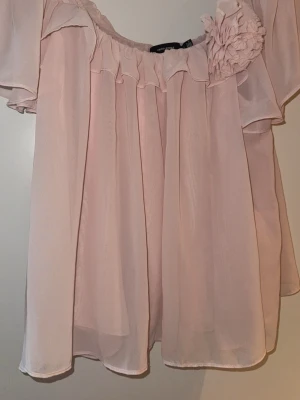 Ljusrosa offshoulder blus med volanger - Supersöt ljusrosa blus i luftig chiffong med offshoulder-modell och korta, vida ärmar. Blusen har volangdetaljer längs ringningen och en stor dekorativ blomma på ena axeln. Perfekt för en romantisk look och passar till både jeans och kjol. Använd 1 gång, storlek L 