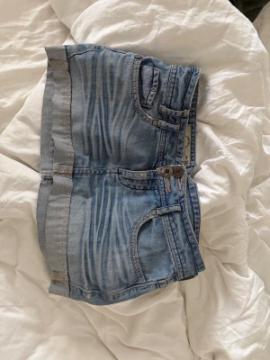 Ljusblå jeansshorts med dubbla knappar - Säljer ett par ljusblå jeansshorts med snygg tvätt och dubbla knappar i midjan. Shortsen har klassiska fem fickor, uppvikta benslut och kontrastsömmar. Perfekta för varma dagar och enkel att matcha med olika stilar.