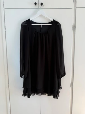 Svart volangklänning med lång ärm - Säljer en svart kortklänning med transparenta långärmar och volangkant nedtill. Klänningen har en lös passform och är tillverkad i ett lätt, flygigt material som ger en snygg siluett. Perfekt för dig som gillar stilrena och enkla plagg med en twist.