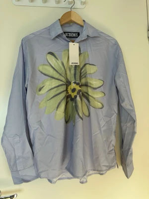 Blå skjorta med blomma från Jacquemus - Säljer en ljusblå skjorta från Jacquemus med en stor blommig print i grönt och svart på framsidan. Skjortan har klassisk krage, långa ärmar och knäppning framtill. Perfekt för dig som vill sticka ut med en unik design.