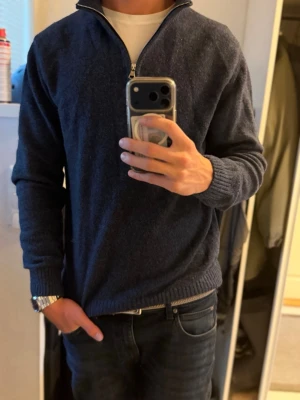 Mörkblå stickad tröja Oscar Jacobson - Mörkblå stickad tröja från Oscar Jacobson med halv dragkedja och hög krage. Tröjan har ribbade muddar vid ärmslut och nederkant, perfekt för lager på lager. Klassisk och stilren design som passar till jeans eller chinos.