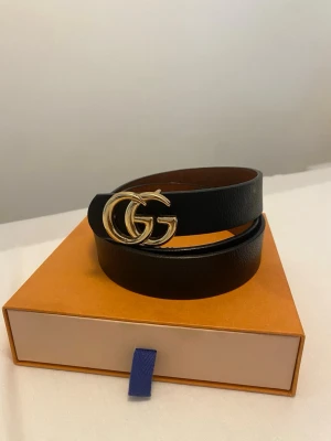 Svart skärp med GG-spänne - Snyggt svart bälte i skinn med ett stort, guldfärgat GG-spänne framtill. Perfekt accessoar för att lyfta vilken outfit som helst. Bältet har en klassisk och stilren design med slät yta och rundade kanter.
