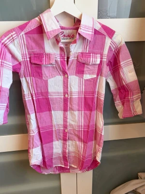 Rosa och vit rutig skjorta - Säljer en rosa och vit rutig skjorta med knappar framtill och två bröstfickor med lock. Skjortan har klassisk krage och trekvartsärmar. Perfekt för dig som gillar färgglada plagg med avslappnad stil. Saknas tre knappar, en vid kragen och de andra två längre ner. Katt finns hemma. 