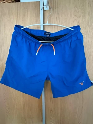 Blå badbyxor från Gant, innerbyxor - Säljer ett par blå badbyxor från Gant med orange och vita snören i midjan. Klassisk modell med resår i midjan och liten Gant-logga broderad på benet. Perfekta för stranden eller poolen. Materialet är snabbtorkande syntet.