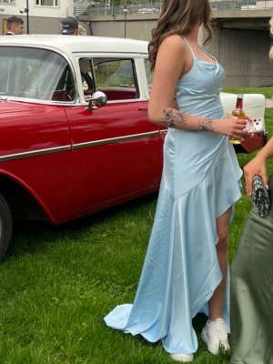 Ljusblå långklänning med slits - Snygg ljusblå långklänning i glansigt material med smala axelband och öppen rygg med snörning. Klänningen har en hög slits framtill och en elegant omlott-design. Perfekt för speciella tillfällen.