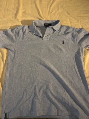 Ljusblå pikétröja från Polo Ralph Lauren - Klassisk ljusblå pikétröja från Polo Ralph Lauren med krage och korta ärmar. Tröjan har den ikoniska broderade loggan på bröstet och är tillverkad i mjuk bomull. Perfekt till en avslappnad men ändå stilren look.