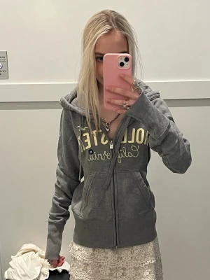 Grå zip hoodie från Hollister - Säljer en grå hoodie från Hollister med dragkedja framtill och tryckt text i vitt och gult på bröstet. Hoodien har huva med snören och två fickor fram. Perfekt för en avslappnad och cool stil.