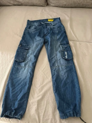 Blå cargobyxor med fickor - Säljer ett par blå cargobyxor i denim med stora fickor på sidorna och broderad text på ena benfickan. Byxorna har avslappnad passform och resår vid benslut. Perfekta för en chill och streetig stil.