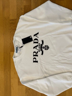 Vit sweatshirt från Prada - Vit sweatshirt från Prada med svart logotyptryck på bröstet och rund halsringning. Tröjan har ribbade muddar vid ärmslut och nederkant. Perfekt för en clean och trendig look.