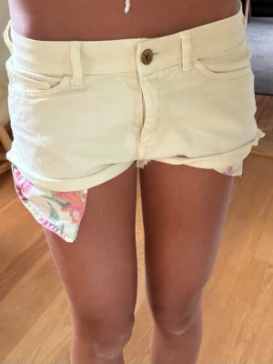 Ljusgula jeansshorts med blommigt foder - Snygga ljusgula jeansshorts med råa kanter och uppvikta ben. Shortsens insida har ett blommigt foder som syns vid uppviket. Klassisk femficksmodell med knapp och dragkedja framtill. Perfekta för sommaren och ger en fräsch look.