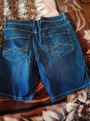 Herr Blå jeansshorts med bakfickor stl 52/L - Säljer ett par klassiska blå jeansshorts med två bakfickor och dekorativa sömmar. Shortsen har bälteshällor och normal passform, perfekta för varma dagar. Tidlös denimstil som funkar till det mesta. Stl 52/L