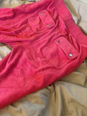 Rosa velourbyxor Juicy Couture - Säljer ett par rosa mjukisbyxor från Juicy Couture i mjuk velour. Byxorna har bred resår i midjan, dragsko med metalländar och fickor med lock och knapp bak. På ena benet finns broderad Juicy Couture-logga. Perfekta för en chill och trendig look.