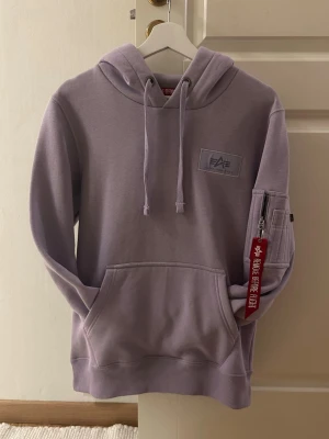 Ljuslila hoodie Alpha Industries - Säljer en ljuslila hoodie från Alpha Industries med stor vit logga på ryggen och mindre logga på bröstet. Hoodien har huva med dragsko, magficka och en ficka med dragkedja på ärmen med röd tag. Mjuk bomullskänsla och ribbade muddar. Storlek XS men kan passar S