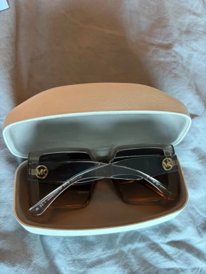 Svarta fyrkantiga solglasögon Michael Kors -  solglasögon från Michael Kors med stora, fyrkantiga genomskinlig bågar och mörka glas. Sidorna har guldfärgade MK-loggor