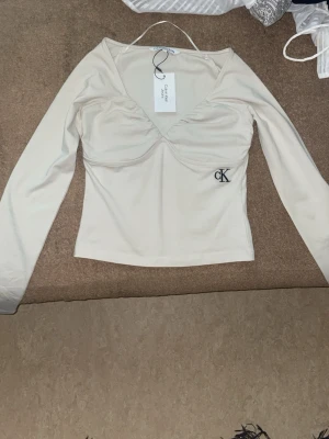 Äkta Vit långärmad topp från Calvin Klein  - Snygg vit långärmad topp från Calvin Klein Jeans med v-ringning och diskret CK-logga broderad på sidan. Toppen har en lätt rynkad detalj vid bysten och är figurnära i modellen. Perfekt att matcha med jeans eller kjol. Jag fick denna i present men den var för liten o för mig den är helt ny alldrig använd nypris 1500kr 