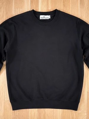 Svart sweatshirt från Stone Island - Klassisk svart sweatshirt från Stone Island med rund halsringning och ribbade muddar vid ärmslut och nederkant. Tröjan har en stilren design och diskret patch på ärmen. Perfekt för en clean och avslappnad look.     Pris går att diskutera vid snabb affär!