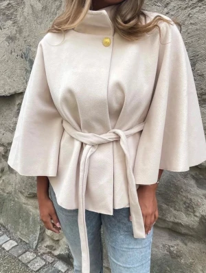 Beige missay poncho med knytband - Stilren beige cape-kappa med vida ärmar, hög krage och ett dekorativt guldknapp vid halsen. Kappan har ett matchande knytband i midjan som ger en snygg siluett. Perfekt för dig som vill ha en trendig och elegant look.
