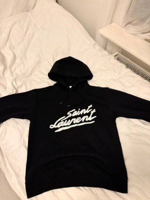 Svart Saint Laurent hoodie med tryck - Svart hoodie från Saint Laurent med stor vit logga i graffiti-stil på bröstet. Klassisk modell med huva och dragsko, samt ribbade muddar vid ärmslut och nederkant. Perfekt för dig som gillar streetwear och vill ha en stilren men ändå statement look.