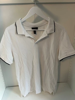 Vit pikétröja från H&M slim fit - Klassisk vit pikétröja från H&M i slim fit-modell. Tröjan har korta ärmar, krage och knappar framtill samt diskreta svarta detaljer på krage och ärmslut. Tillverkad i bomull för en bekväm känsla.