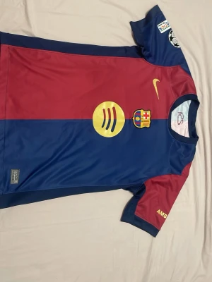 FC Barcelona hemma matchtröja Lamine Yamal 24/25 - Snygg FC Barcelona matchtröja i rött och blått med guldiga detaljer, klubbmärke och Nike-logga på bröstet. Nummer 19 och Lamine Yamal på ryggen. Korta ärmar, flera tryck och märken, tillverkad i funktionsmaterial som passar perfekt till fotbollsträning.
