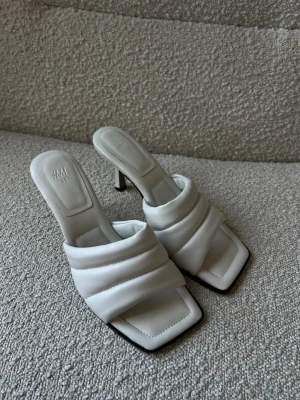 Vita slip-in klackar H&M - Säljer ett par vita pumps från H&M med quiltad design och öppen häl. Skorna har fyrkantig tå, smal klack och breda vadderade remmar i syntetmaterial. Perfekta för dig som gillar trendiga och stilrena skor med en modern twist.