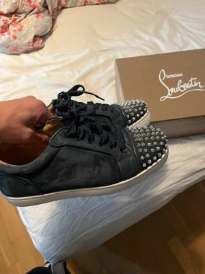 Mörkblå louboutin skor med silvriga nitar - Unika mörkblå sneakers i mocka med coola silverfärgade nitar på tån. Skorna har mörkblå snören och vit sula. Insidan är beige och materialet känns lyxigt. Perfekta för dig som vill sticka ut med din stil.