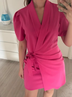 Rosa omlottklänning med knyt - Säljer en kort rosa omlottklänning med puffiga korta ärmar och knyt i midjan. Klänningen har en v-ringad omlottfront och en figurnära passform som ger en snygg siluett. Perfekt för dig som vill sticka ut med färg och stil.