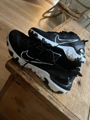 Svarta Nike reacts - Snygga svarta Nike sneakers med vit sula och tydlig vit swoosh på sidan. Ovandelen är i mesh och syntetmaterial för bra andningsförmåga. Sulan har ett unikt mönster för extra grepp och cool look. Perfekta för dig som vill ha stilrena och sportiga skor.