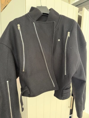 Helt ny Svart bikerjacka från Zara - Snygg svart bikerjacka från Zara med flera silverfärgade dragkedjor framtill och på ärmarna. Jackan har en ribbad hög krage och kort, boxig passform. Materialet är kraftigt och ger en cool look. Perfekt för dig som vill ha en edgy stil.