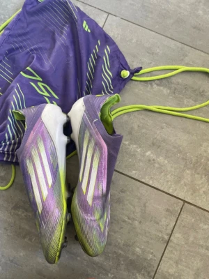 Lila Adidas F50 fotbollsskor - Säljer ett par lila Adidas F50 fotbollsskor med vita och neongula detaljer. Skorna har en strömlinjeformad design och fasta dobbar för bra grepp på planen. Medföljer matchande lila och neongul skopåse. Använda i några månader under vintersäsongen då vi även spelade innomhus 