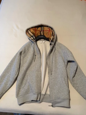 Grå hoodie med mönstrad huva - Säljer en stilren grå hoodie med dragkedja och fickor. Huvan har ett klassiskt rutigt mönster på insidan som ger en snygg kontrast. Tillverkad i mjukt sweatshirtmaterial, perfekt för en avslappnad look. Dragsko i huvan för justerbar passform.