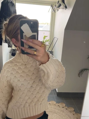 Krämvit stickad tröja med struktur - Säljer en krämvit stickad tröja med hög hals och ballongärmar. Tröjan har ett snyggt strukturmönster och breda ribbade muddar vid ärmslut och nederkant. Perfekt för dig som gillar en mysig och trendig look.