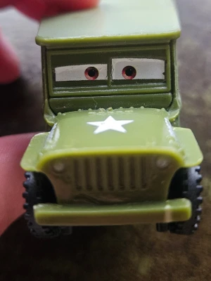 Grön leksaksbil med ögon - Säljer en grön leksaksbil i plast som liknar en militärjeep med vita detaljer och en stjärna på motorhuven. Bilen har tecknade ögon på vindrutan och svarta hjul med tydligt mönster. Perfekt för samlare eller som leksak.