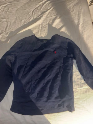 Mörkblå sweatshirt från Polo Ralph Lauren - Mörkblå sweatshirt från Polo Ralph Lauren med rund halsringning och den klassiska röda logotypen broderad på bröstet. Tröjan har långa ärmar och mjukt material som passar perfekt till en avslappnad stil.