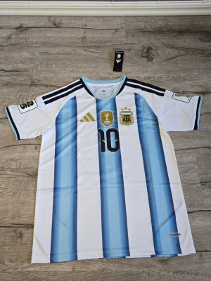 Argentina fotbollströja Messi #10 - Snygg Argentina landslagströja från Adidas med Messis namn och nummer 10 på ryggen. Tröjan är vit med ljusblå ränder, svarta detaljer och guldiga logotyper. Perfekt för fotbollsfans som vill visa sitt stöd för Messi och Argentina.