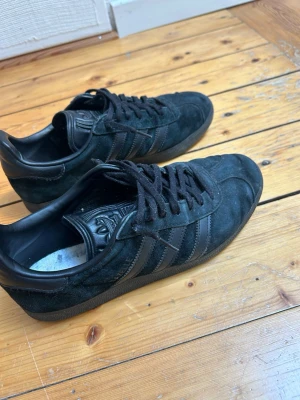 Svarta Adidas sneakers i mocka - Säljer ett par svarta Adidas Gazelle sneakers i mocka med klassiska tre ränder på sidan och Adidas-logga på plösen. Skorna har svarta snören och en låg profil som ger en clean look. Perfekta för dig som gillar stilrena och tidlösa sneakers. Storlek 42 och köpta på adidas. Sulan är tyvärr borta och hittar inte den.
