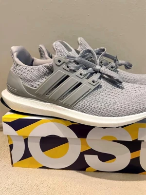 Grå Adidas Ultra Boost sneakers - Snygga grå Adidas Ultra Boost sneakers med stickad ovandel i mesh för extra andningsförmåga. Skorna har klassiska tre ränder på sidan, vit Boost-mellansula och svart yttersula från Continental. Perfekta för dig som gillar stilrena och bekväma sneakers.