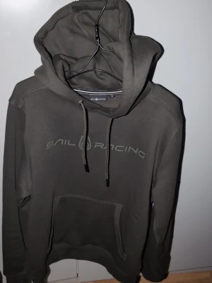 Mörkgrön hoodie från Sail Racing - Snygg mörkgrön hoodie från Sail Racing med stor huva och dragsnören. Hoodien har en klassisk känguruficka framtill och logga tryckt över bröstet. Tillverkad i mjukt bomullsmaterial som känns skönt mot huden. Perfekt för en avslappnad och sportig stil. Helt ny skulle jag säga haft på mig den en timme på stan en gång så den ser exakt som ny ut :) ordinarie pris 1499 säljes för 799
